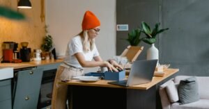 A young woman unpacks online shopping orders in a stylish modern kitchen, using her laptop for work.