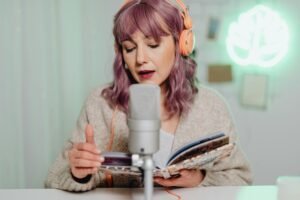 Woman wears headphones and records audio while reading from a book in a cozy setting.